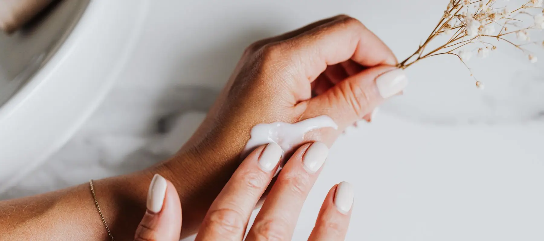 Mains appliquant une crème de soin, un brin de gypsophile séchée entre les doigts